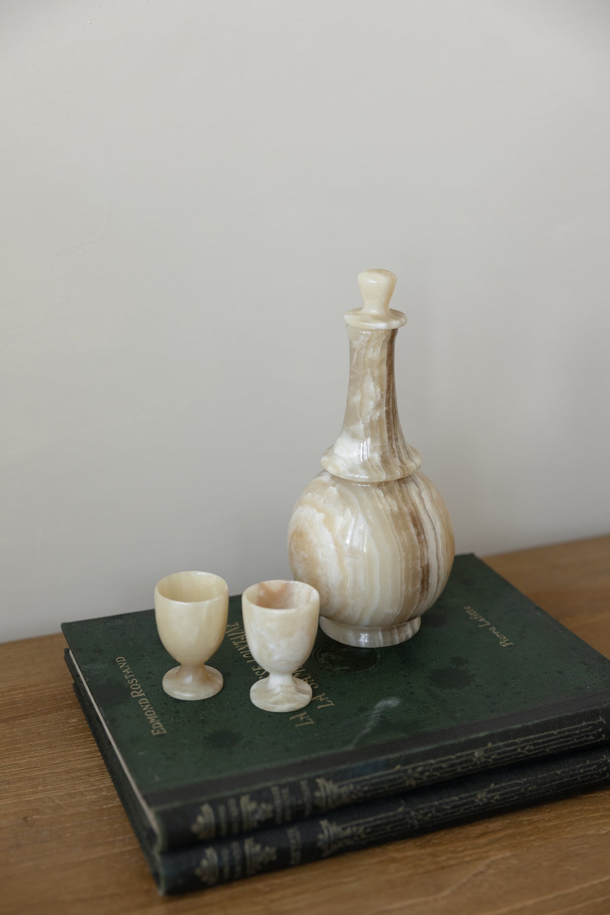 A decorative carved stone or bone bottle with a stopper, displayed on top of two dark green vintage books next to two matching small cups on a wooden surface against a plain light gray wall.