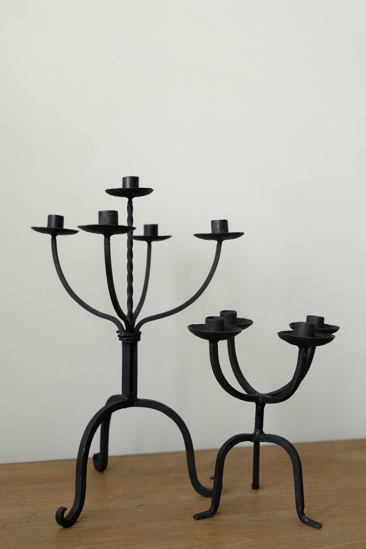 Two black wrought iron candlesticks, one five-arm and one three-arm, displayed on a wooden surface against a plain off-white wall.