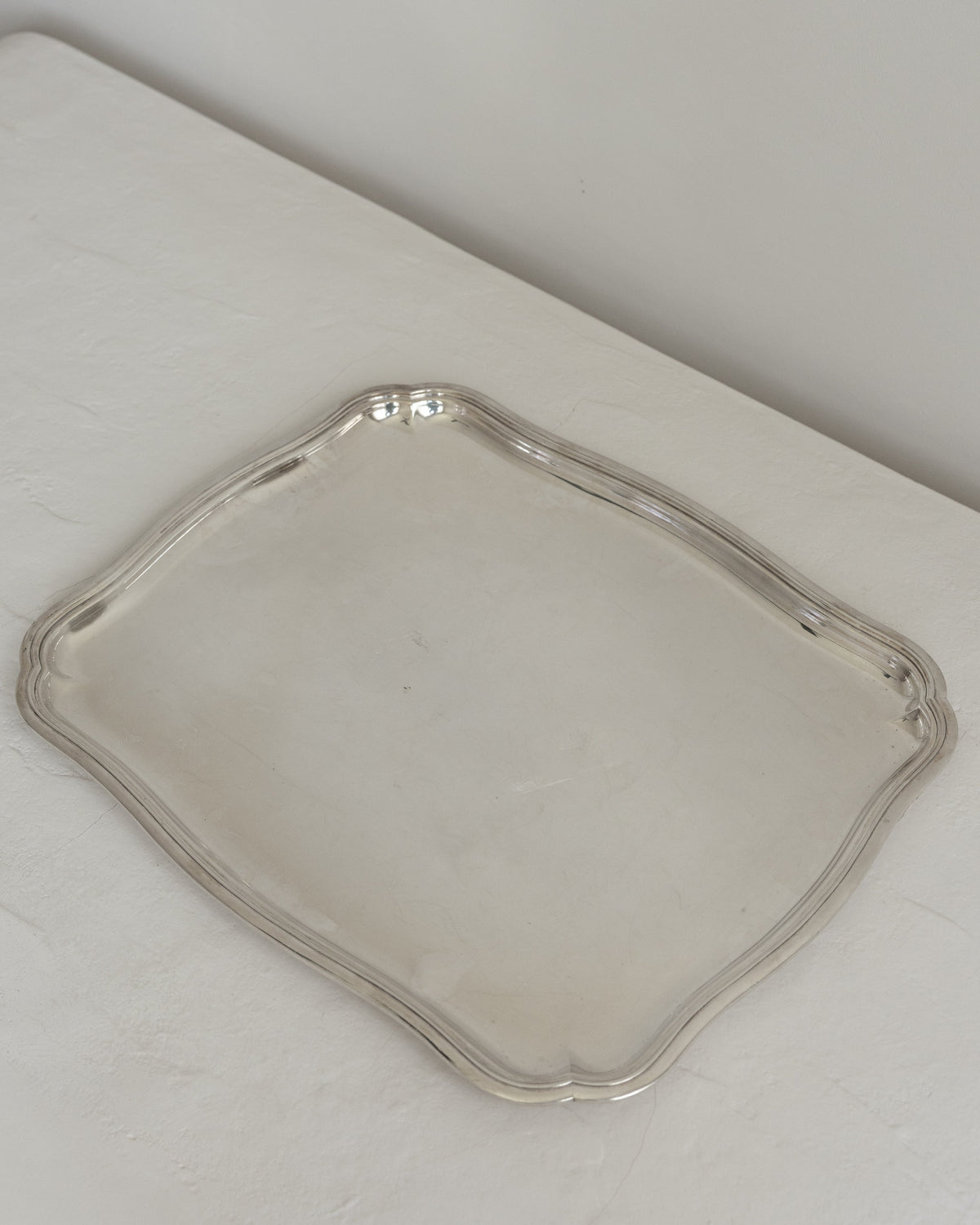 A square silver serving tray with scalloped edges, displayed on a white textured surface against a neutral light gray background.
