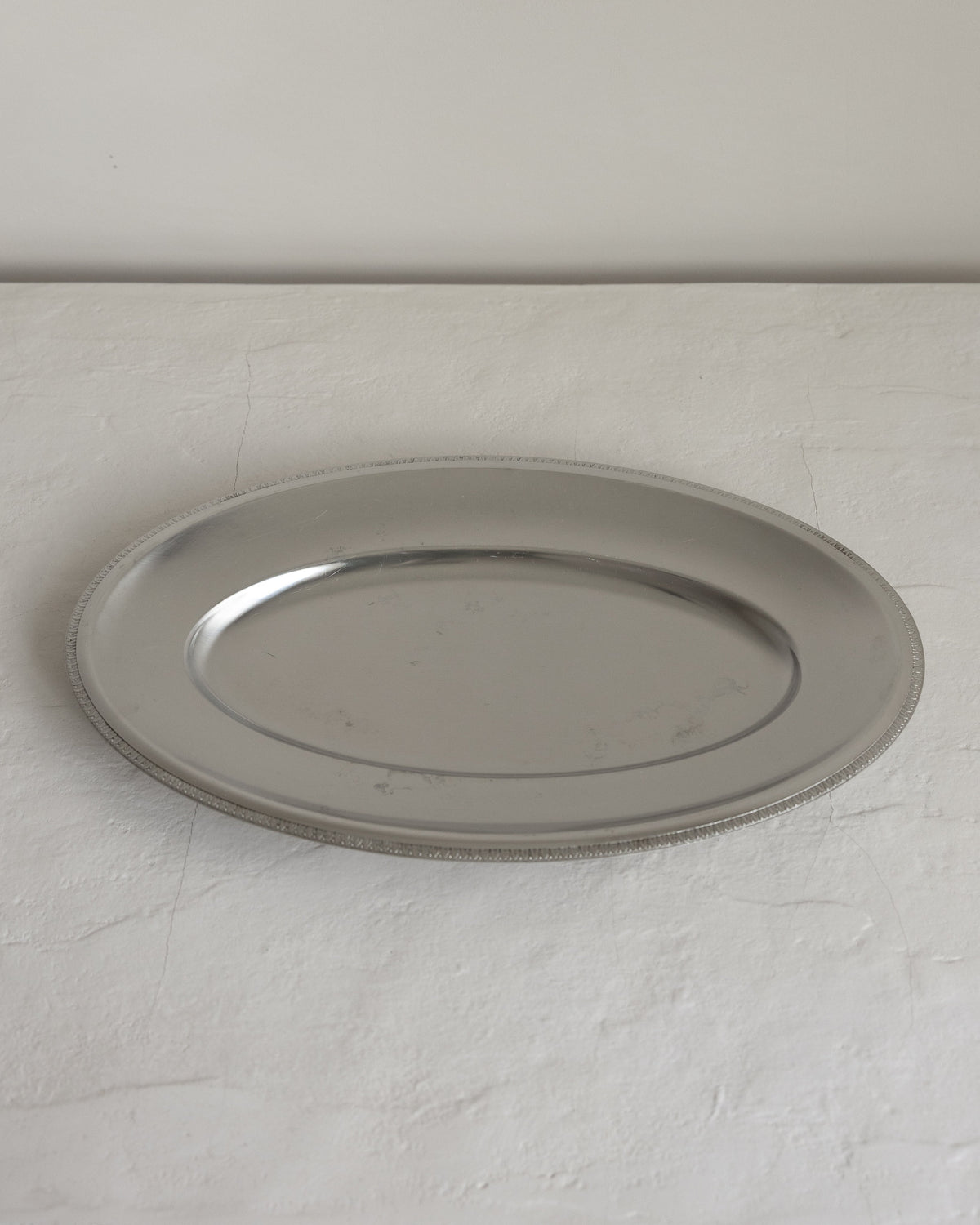 A round silver metal serving platter with a raised rim, photographed on a light textured surface against a plain light gray wall.