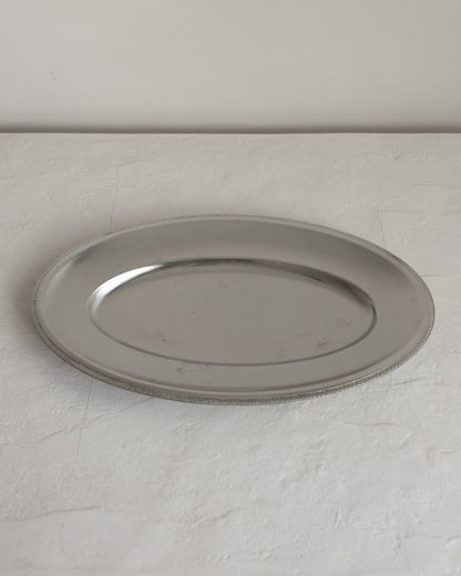 A round silver metal serving platter with a raised rim, photographed on a light textured surface against a plain light gray wall.