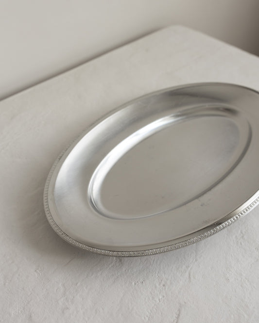 An oval silver serving platter with a beaded rim, shown on a textured off-white surface against a neutral light gray background.