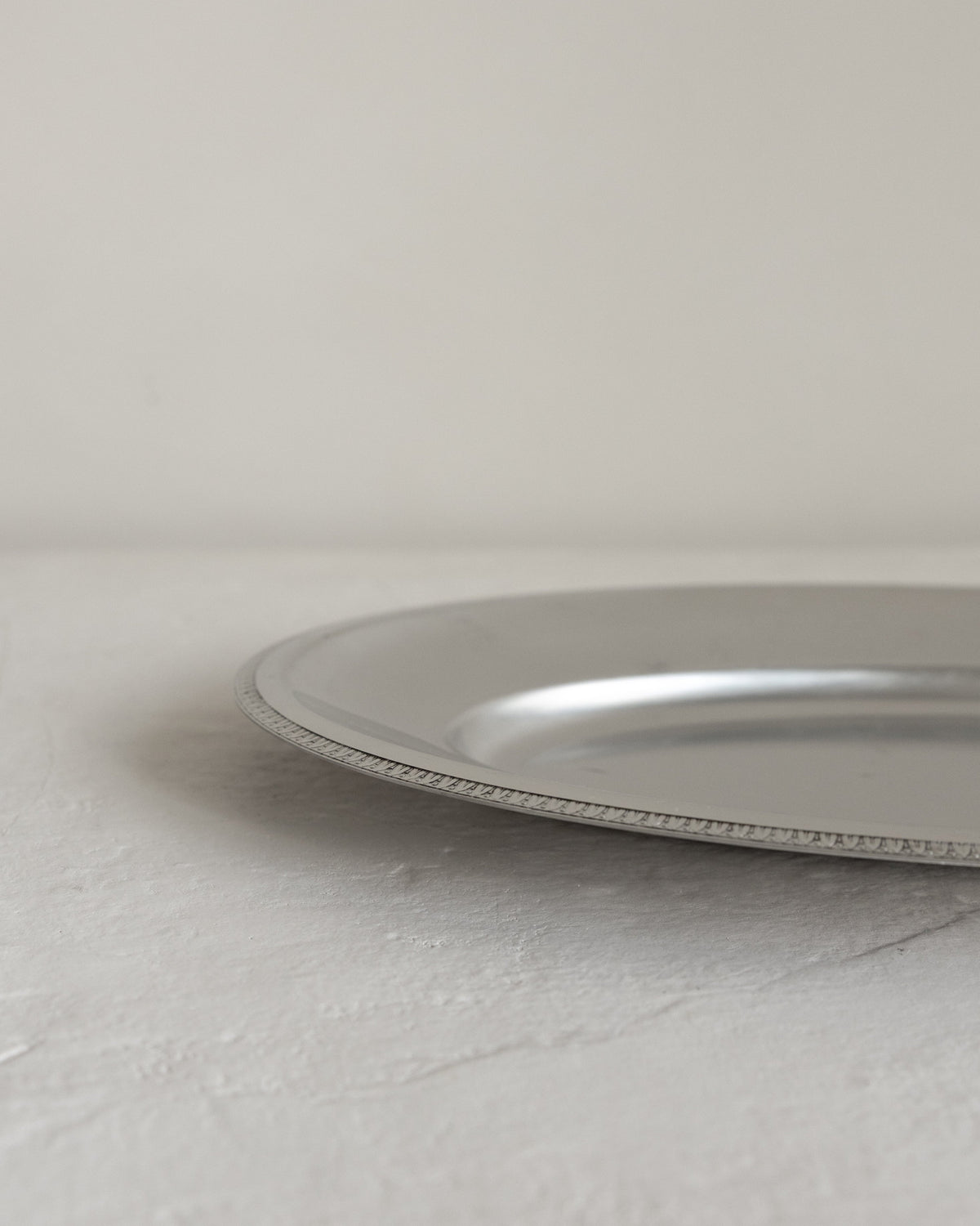 A close-up of a silver metal tray or shallow dish with a textured beaded rim, resting on a light gray stone surface against a neutral off-white background.