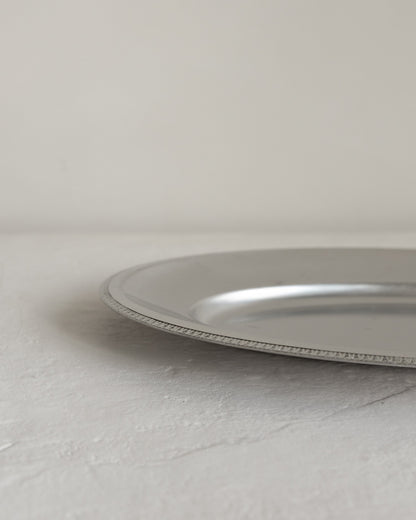 A close-up of a silver metal tray or shallow dish with a textured beaded rim, resting on a light gray stone surface against a neutral off-white background.