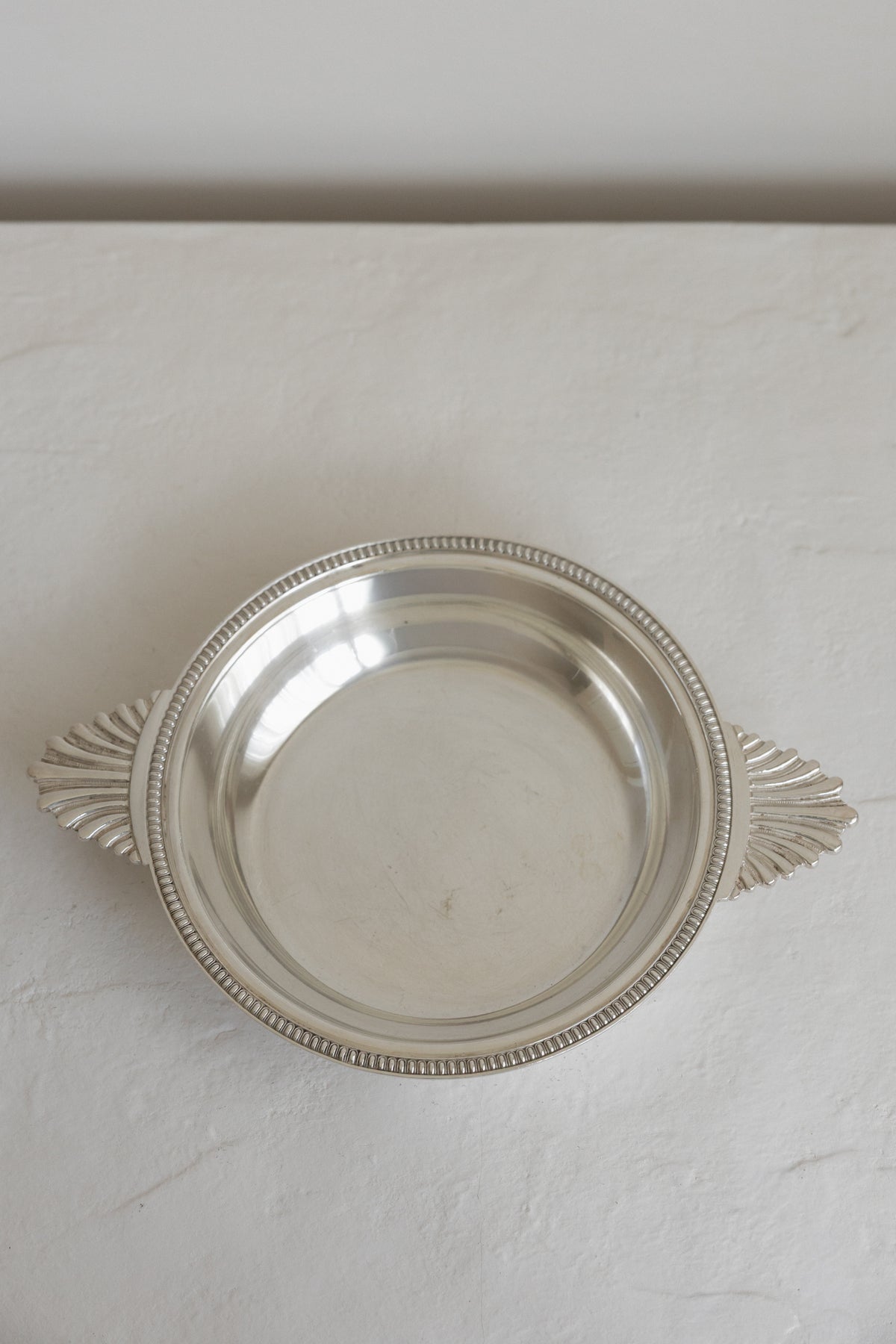 A round silver serving platter with decorative leaf-shaped handles and a beaded rim, shown on a light textured surface.