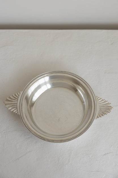 A round silver serving platter with decorative leaf-shaped handles and a beaded rim, shown on a light textured surface.