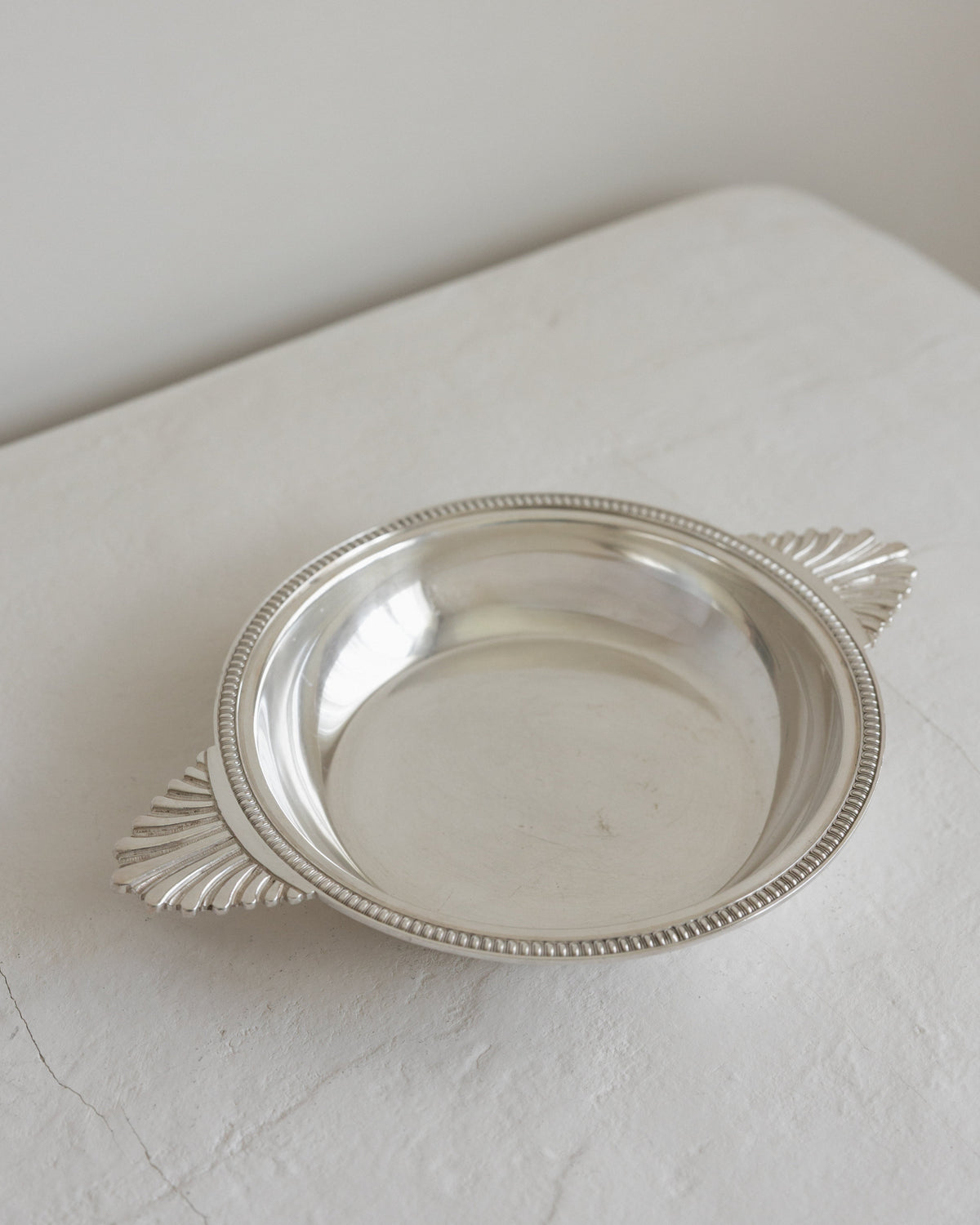 A small oval silver tray with decorative leaf-like side handles and a beaded rim, photographed on a light fabric surface against a plain off-white background.