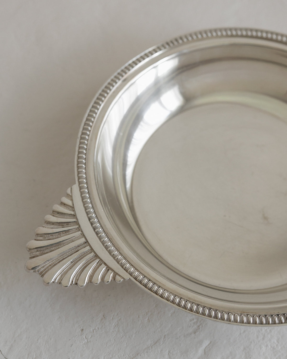 A close-up of a polished silver metal dish with a decorative beaded rim and a fan-shaped handle, resting on a textured off-white surface.