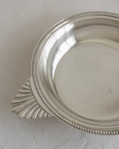A close-up of a polished silver metal dish with a decorative beaded rim and a fan-shaped handle, resting on a textured off-white surface.