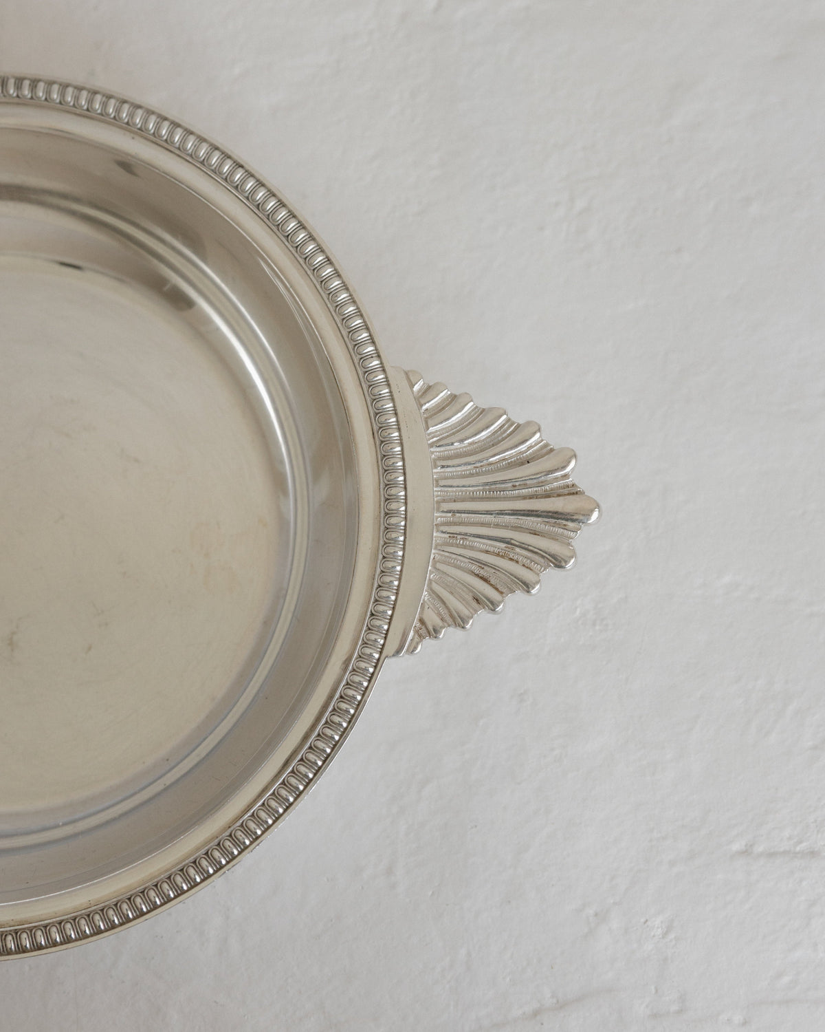 A close-up of a silver metal dish or bowl with a decorative beaded rim and a fan-shaped handle, photographed on a textured off-white surface.