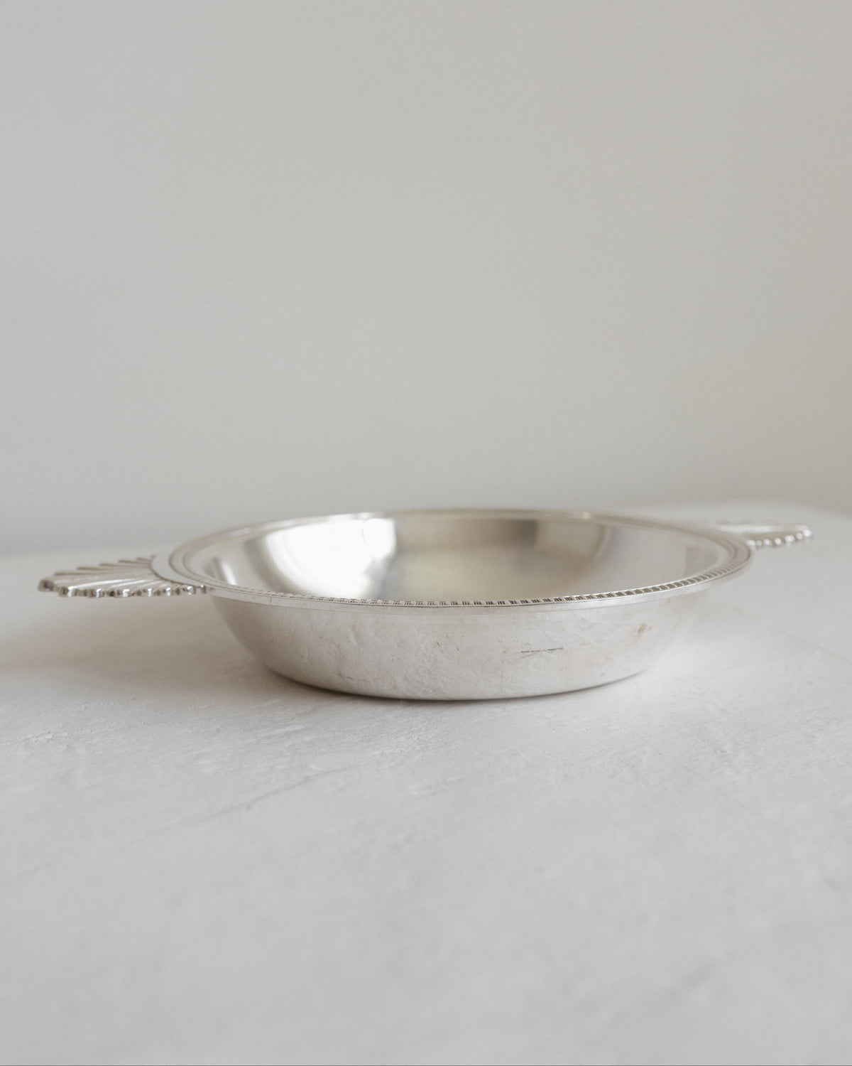A small silver metal bowl with loop handles, photographed on a light neutral surface against a soft light gray background.