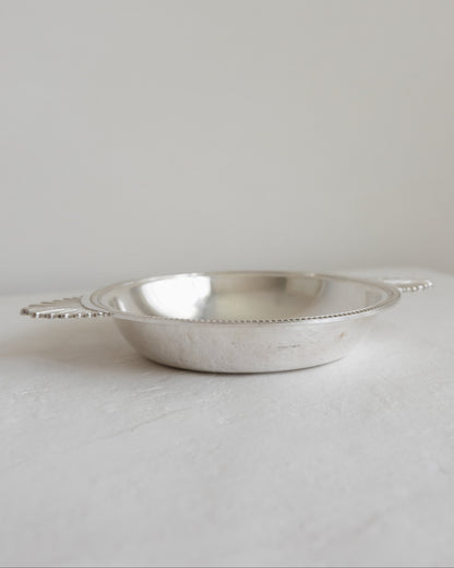 A small silver metal bowl with loop handles, photographed on a light neutral surface against a soft light gray background.