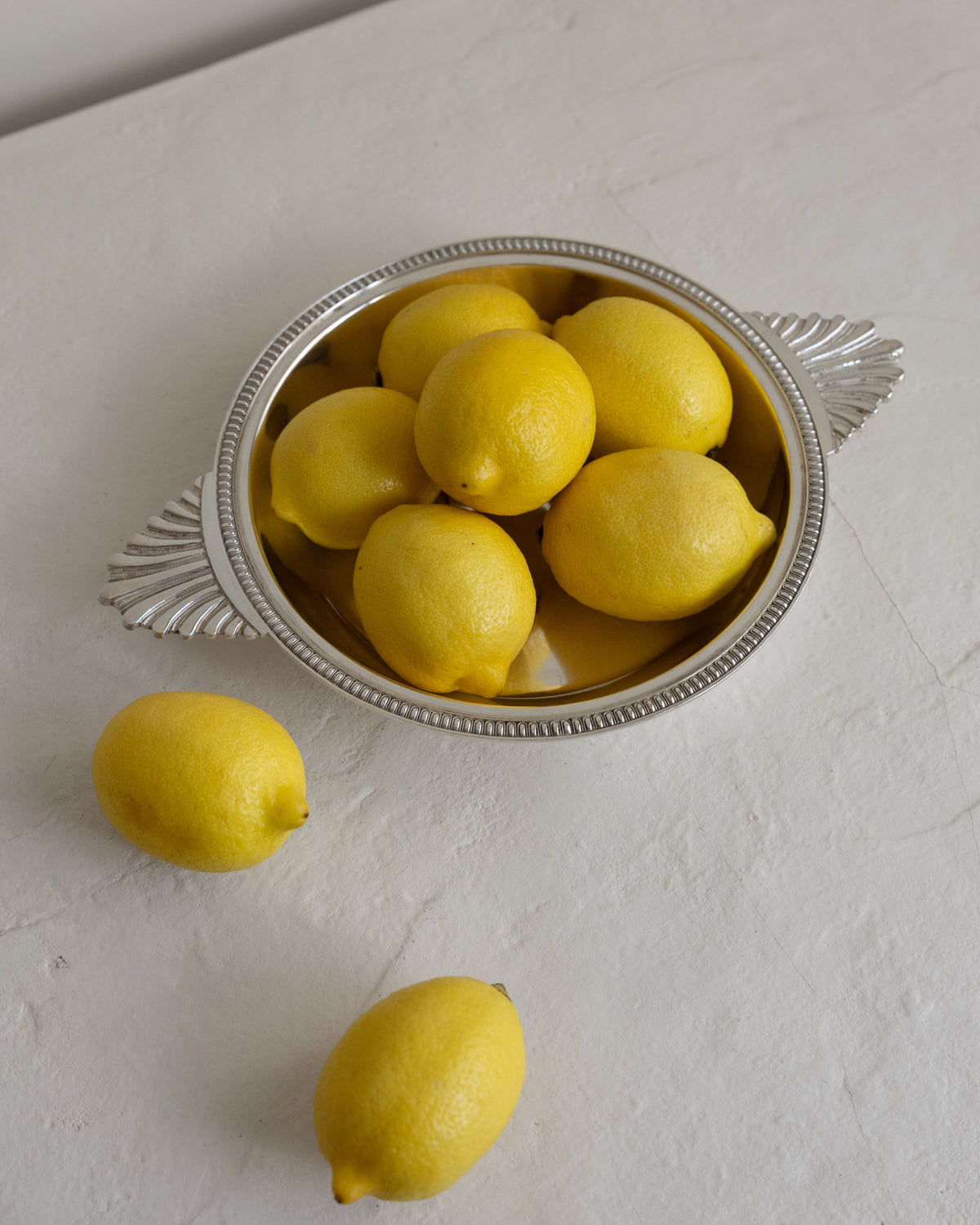 Several bright yellow lemons arranged in a silver bowl with decorative side handles, with two lemons placed outside the bowl on a light textured surface.