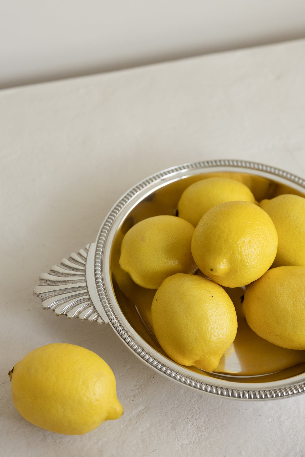 Several bright yellow lemons arranged in a silver bowl with a decorative handle, with one lemon placed outside the bowl on a light neutral surface.