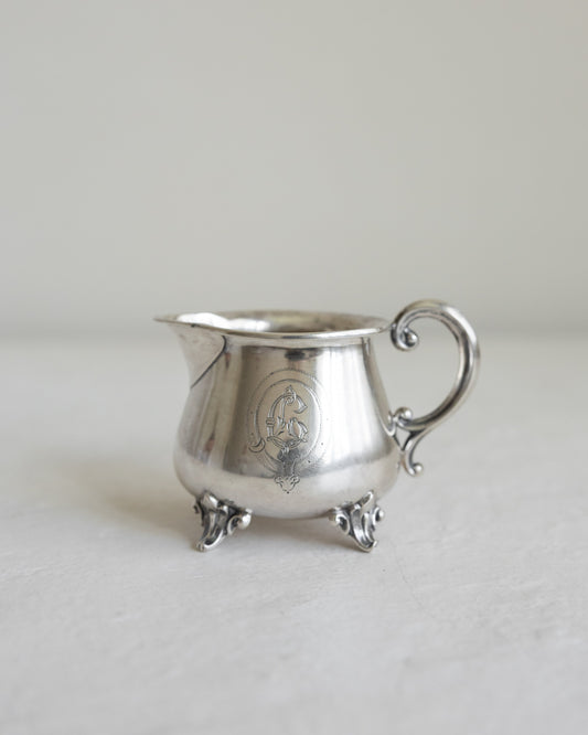A small silver creamer pitcher with ornate feet, a curved handle, and an engraved crest on the front, set against a neutral light gray background.