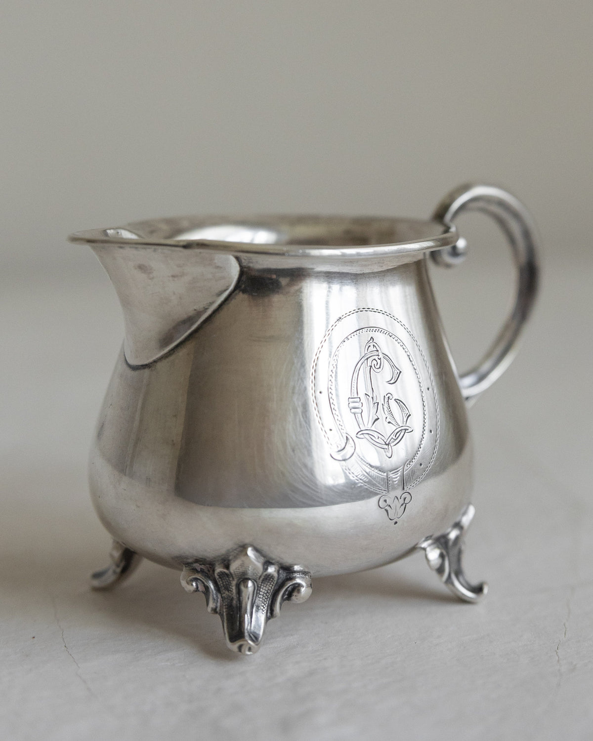 A vintage sterling silver creamer pitcher with ornate scrollwork feet, a curved handle, and an engraved crest on the side, set against a neutral light gray background.
