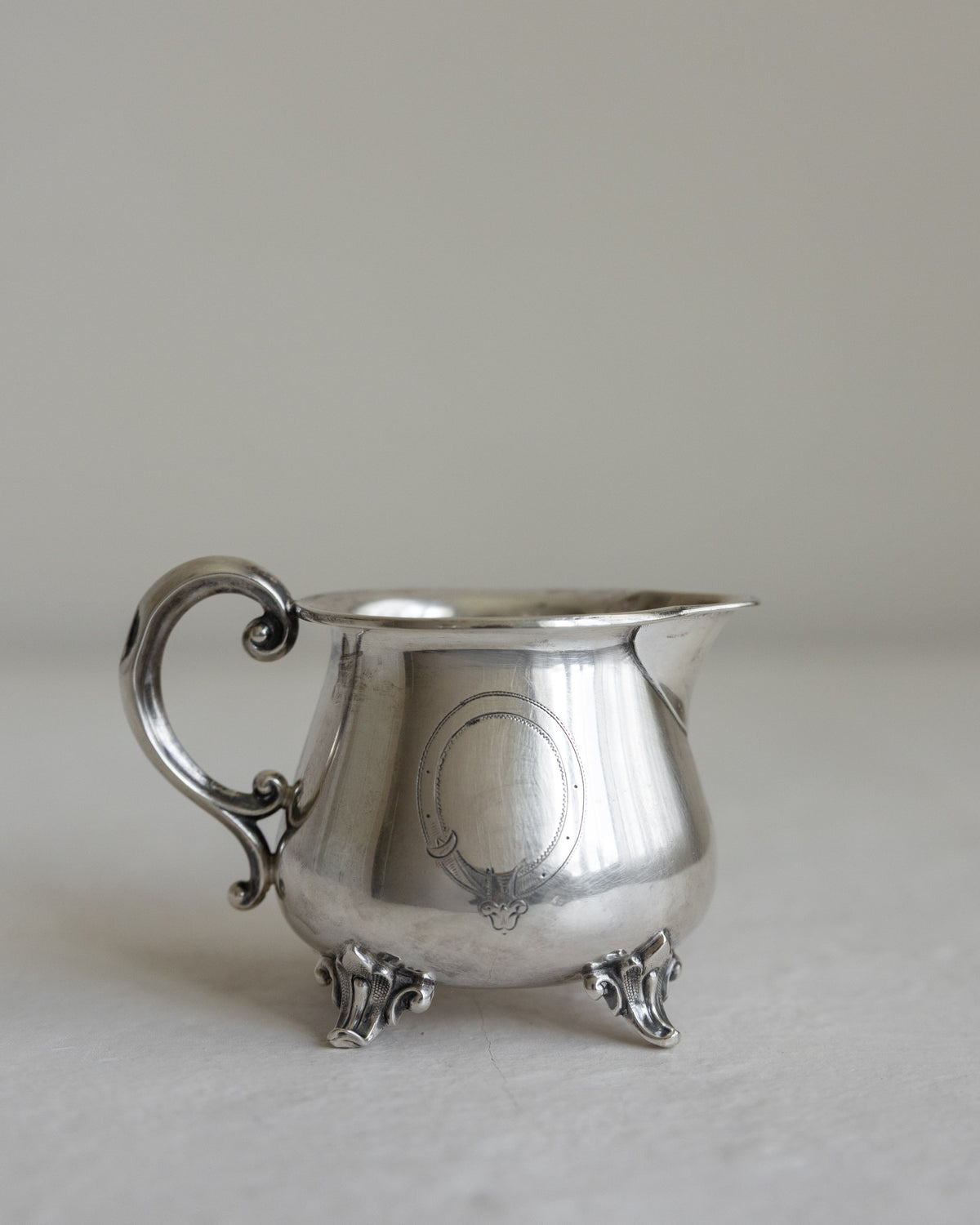 A small silver creamer or milk pitcher with an ornate scroll handle and three feet, shown on a neutral light gray background.