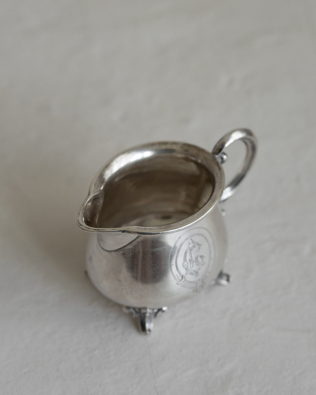 A small silver creamer or milk pitcher with a loop handle and three feet, photographed on a light textured surface.