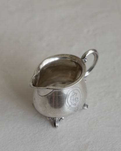 A small silver creamer or milk pitcher with a loop handle and three feet, photographed on a light textured surface.