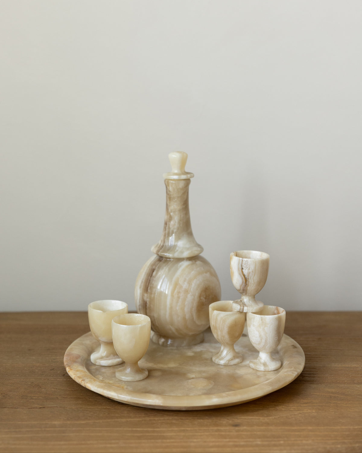 A set of carved stone drinking vessels on a round matching tray, including a decanter and four small cups, displayed on a wooden surface against a plain light gray wall.