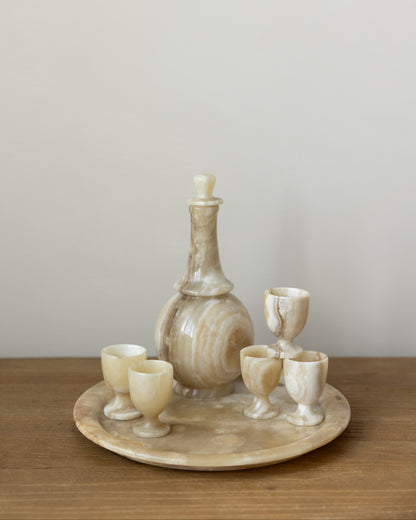 A set of carved stone drinking vessels on a round matching tray, including a decanter and four small cups, displayed on a wooden surface against a plain light gray wall.