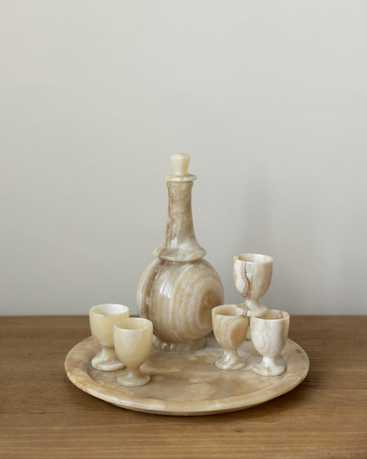 A set of carved stone drinking vessels on a round matching tray, including a decanter and four small cups, displayed on a wooden surface against a plain light gray wall.