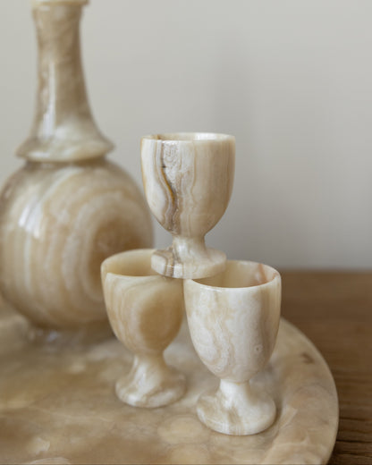 A set of polished onyx stone drinking vessels, including a decanter and two goblets, arranged on a matching stone tray against a soft neutral background.