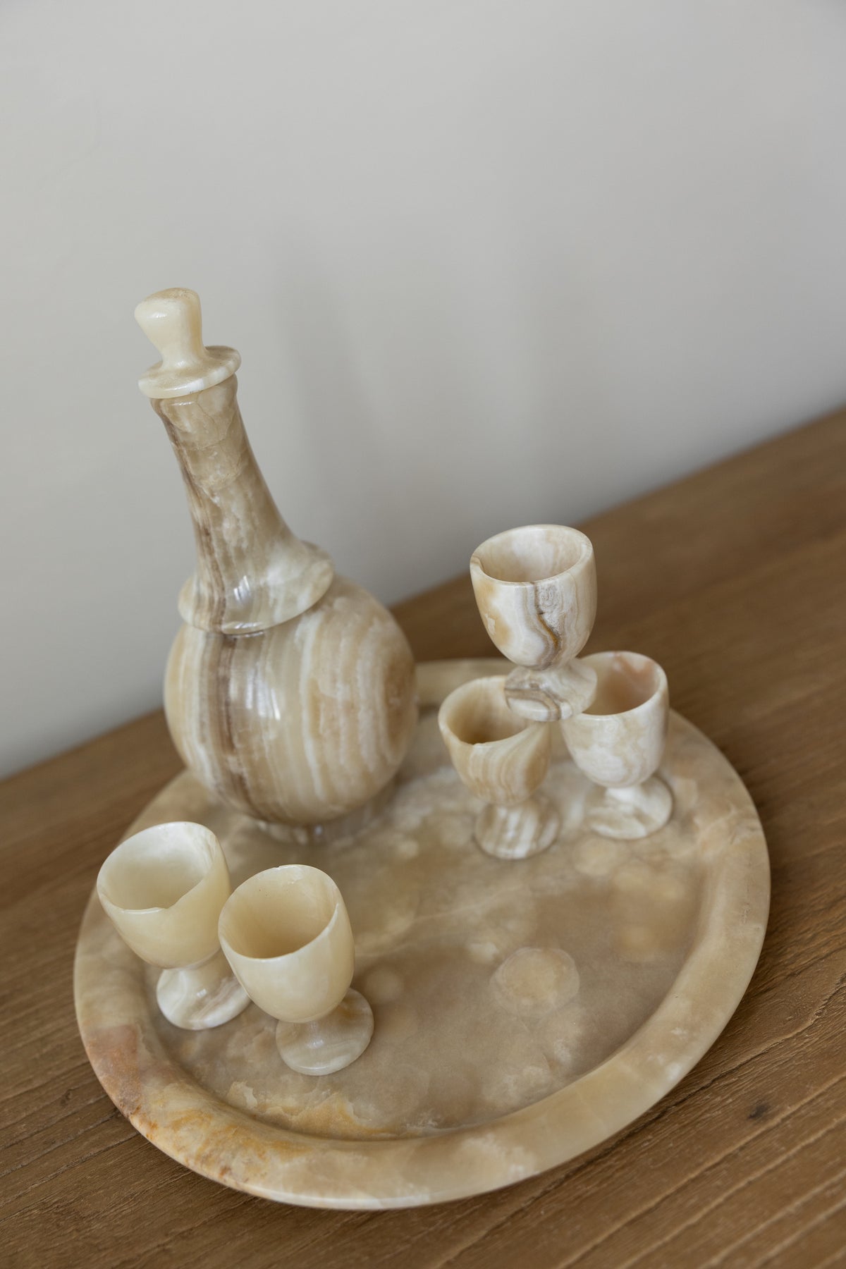 A carved stone or alabaster beverage set on a round matching tray, including a decanter and four small cups, photographed on a wooden tabletop against a plain light wall.