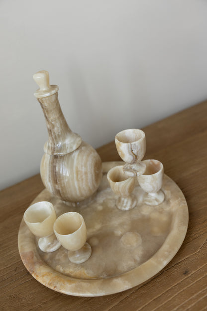 A carved stone or alabaster beverage set on a round matching tray, including a decanter and four small cups, photographed on a wooden tabletop against a plain light wall.