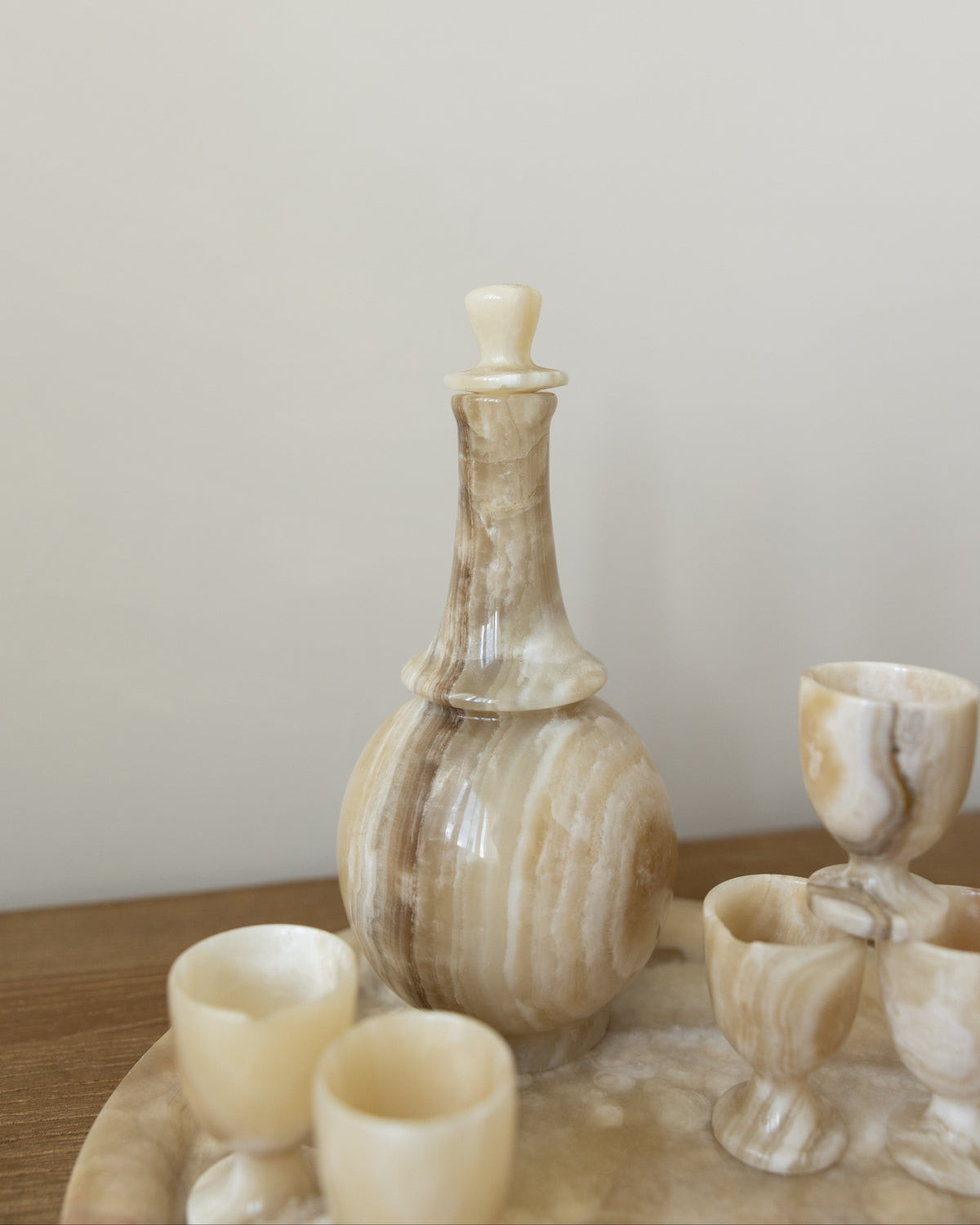 A set of carved stone drinking vessels, including a tall bottle with a stopper and several small cups, arranged on a wooden tray against a plain light wall.