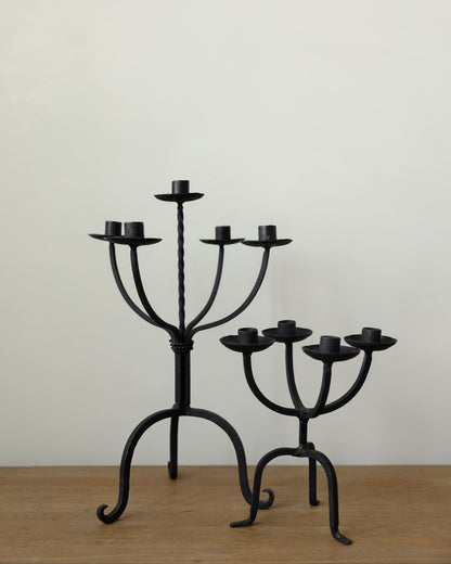 Two black wrought iron candelabras on a light wood surface against a plain off-white wall.