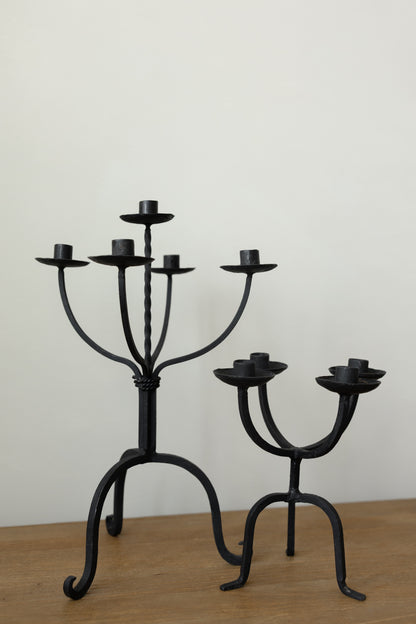 Two black wrought iron candlesticks, one five-arm and one three-arm, displayed on a wooden surface against a plain off-white wall.