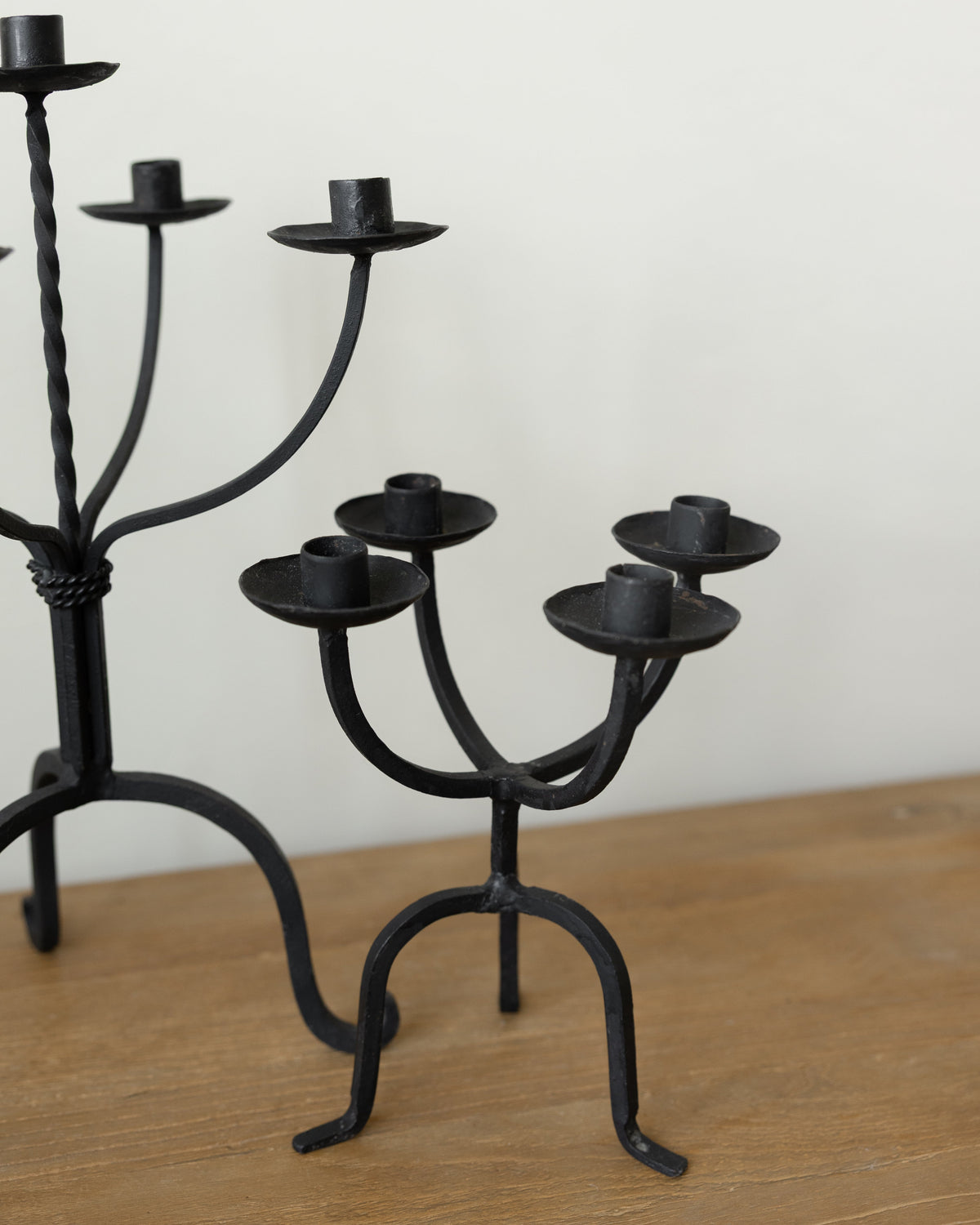 Black wrought iron candlestick holders displayed on a wooden tabletop against a plain off-white wall.