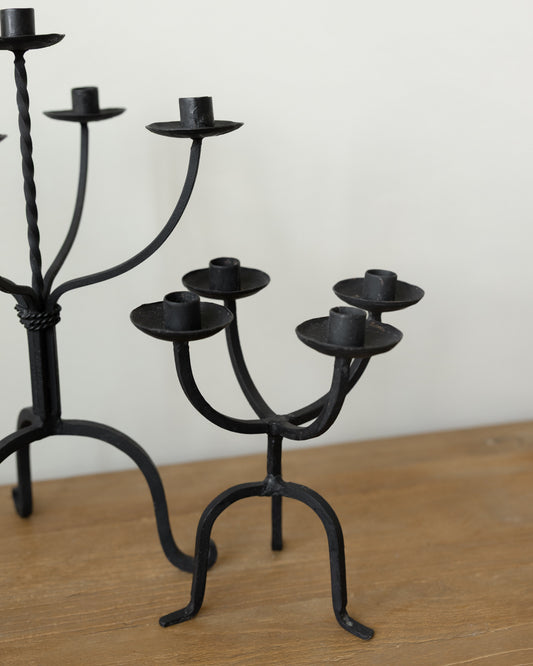 Black wrought iron candlestick holders displayed on a wooden tabletop against a plain off-white wall.