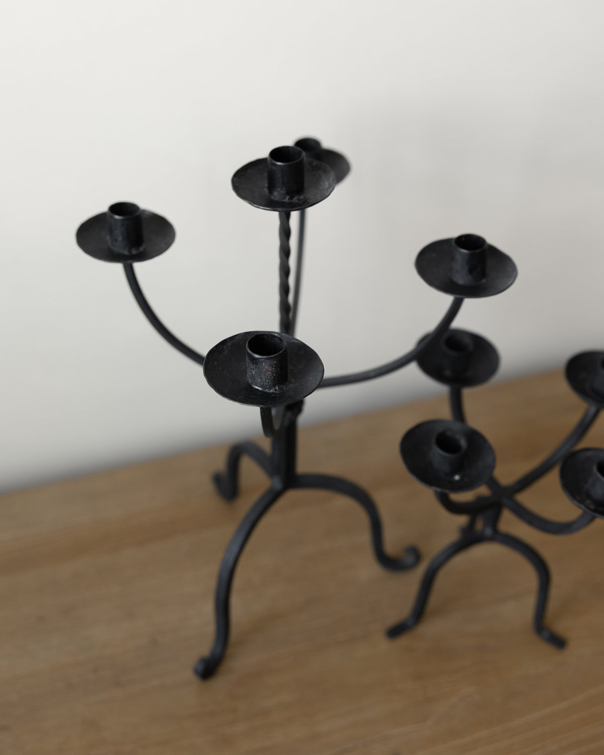 Black wrought iron candelabra candle holders displayed on a light wood tabletop against a plain off-white wall.