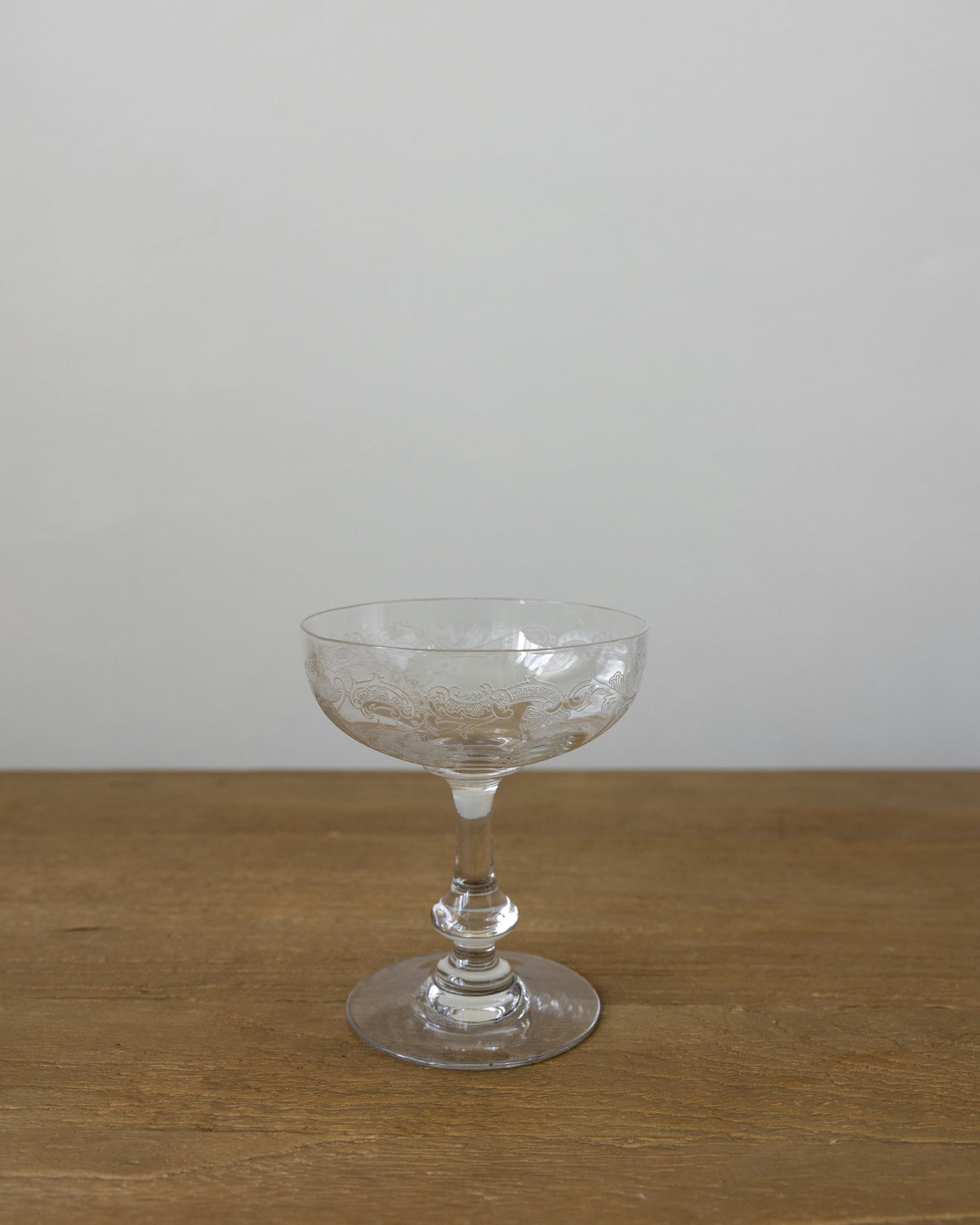 A clear glass coupe-shaped cocktail glass with a textured pattern on the bowl, standing on a wooden surface against a plain light gray wall.