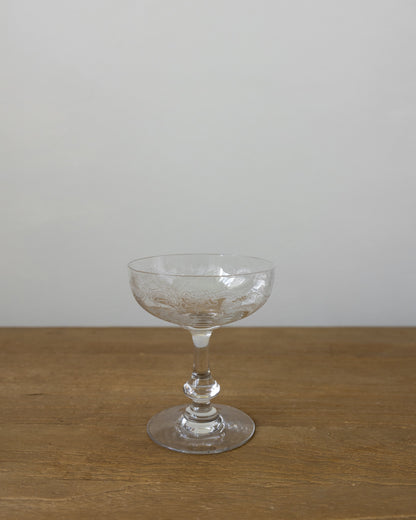 A clear glass coupe-shaped cocktail glass with a textured pattern on the bowl, standing on a wooden surface against a plain light gray wall.