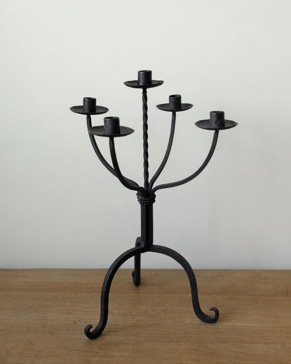 A black wrought iron five-arm candelabra with a twisted central stem and curved scroll-style legs, standing on a wooden surface against a plain off-white wall.