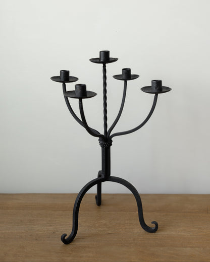 A black wrought iron five-arm candelabra with a twisted central stem and curved scroll-style legs, standing on a wooden surface against a plain off-white wall.