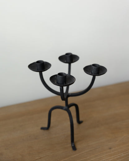 A black metal four-arm candelabra candle holder with a rustic, hand-forged look, standing on a light brown wooden surface against a plain off-white wall.