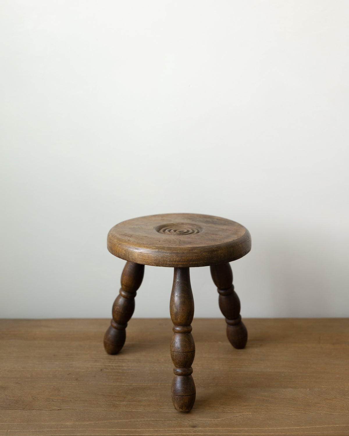 A small round wooden stool with four turned legs, shown on a wooden floor against a plain off-white wall.