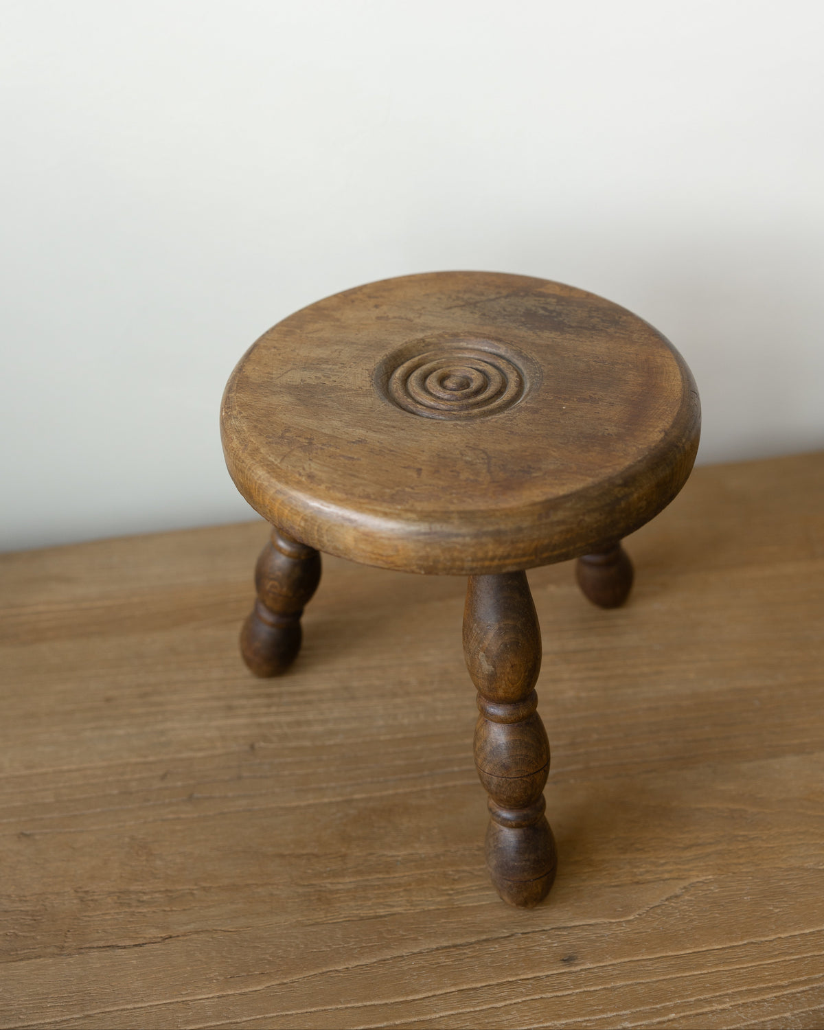A small round wooden stool with turned legs and a carved spiral detail on the seat, shown on a wooden surface against a plain light wall.