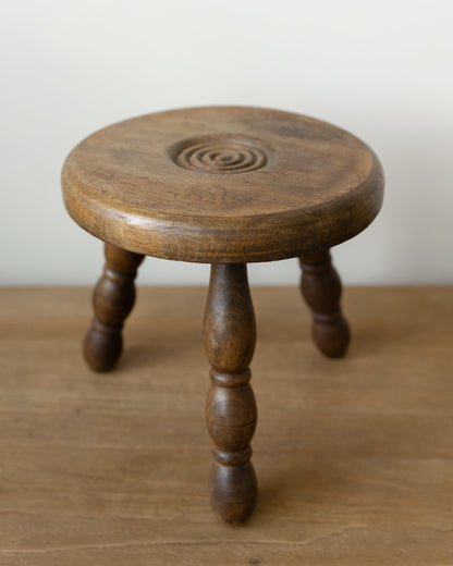 A small round wooden stool with four turned legs and a carved spiral detail on the seat, shown on a wooden surface against a plain light background.
