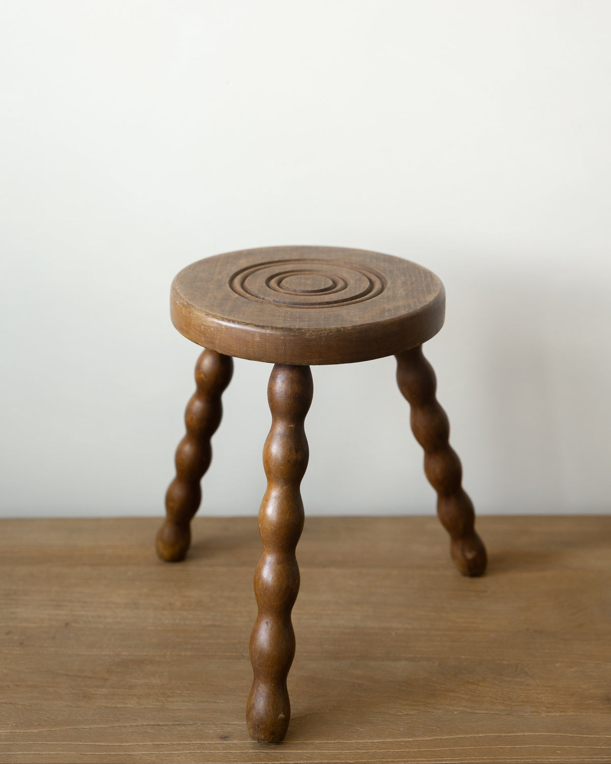 A small round wooden stool with three twisted legs, shown on a wooden surface against a plain off-white wall.