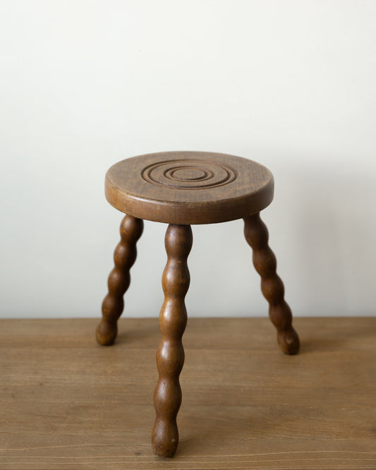 A small round wooden stool with three twisted legs, shown on a wooden surface against a plain off-white wall.