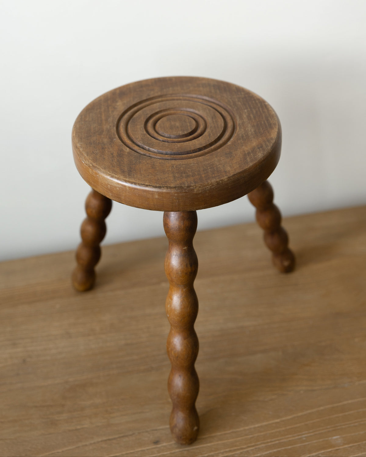A small round wooden stool with a carved circular pattern on the seat and turned spindle-style legs, shown on a wooden surface against a plain light wall.