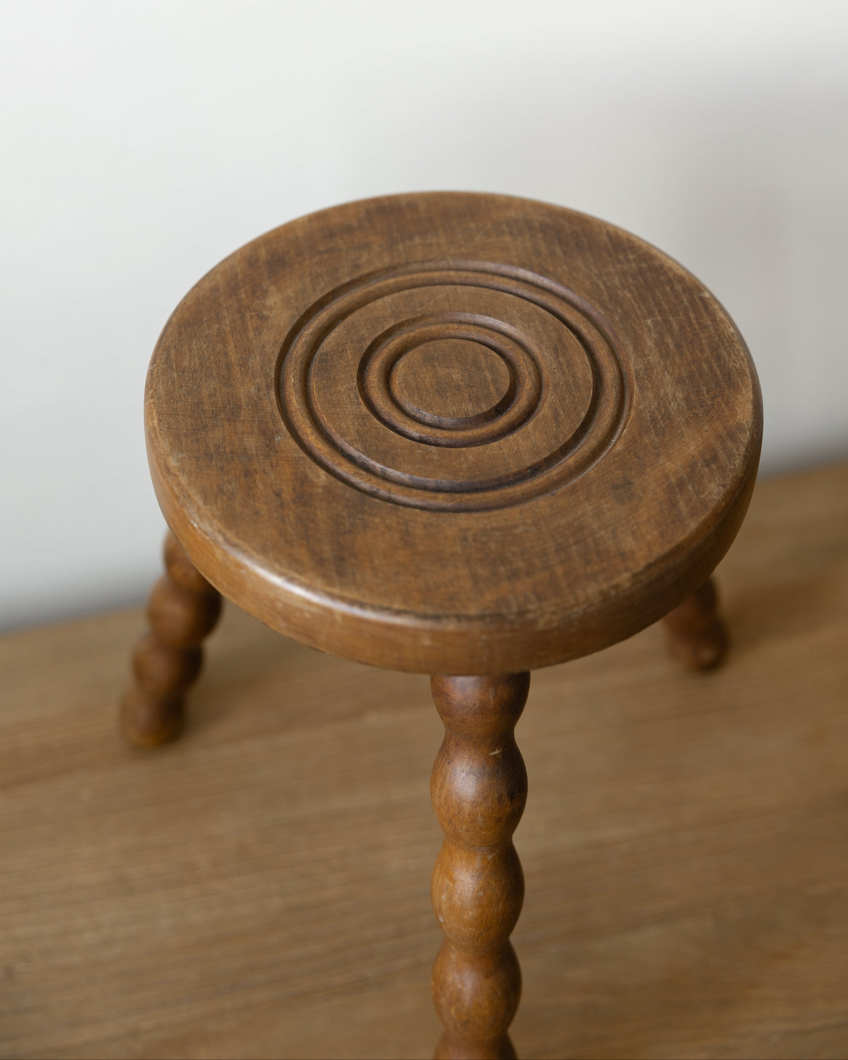 A small round wooden stool with a carved concentric circle pattern on the seat and turned legs, shown on a wooden surface against a plain light background.