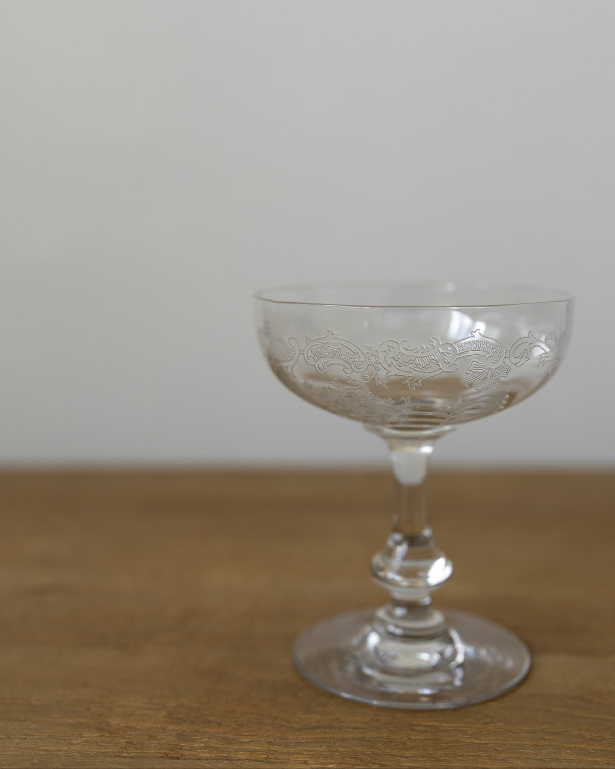 A clear glass coupe-shaped cocktail glass with subtle etched floral detailing, resting on a wooden surface against a plain light gray wall.