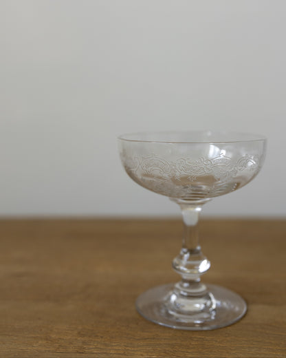 A clear glass coupe-shaped cocktail glass with subtle etched floral detailing, resting on a wooden surface against a plain light gray wall.