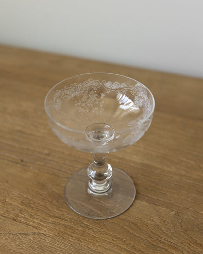 A clear glass coupe-shaped dessert goblet with etched floral detailing, resting on a wooden tabletop against a neutral light gray background.